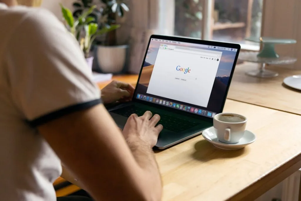 Osoba korzystająca z laptopa z otwartą stroną główną Google, siedząca przy drewnianym stole z filiżanką kawy i rośliną w tle.