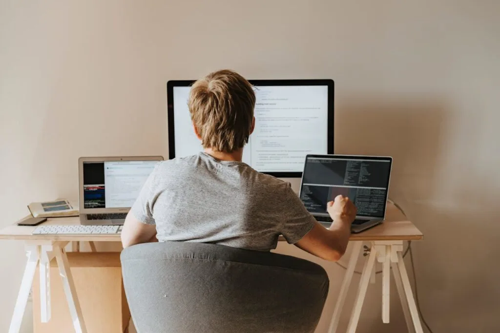 Mężczyzna siedzi przy biurku, korzystając z dwóch laptopów, skupiony na pracy w domowym biurze.